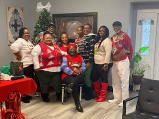 Group of people in festive holiday sweaters posing together near a decorated Christmas tree.